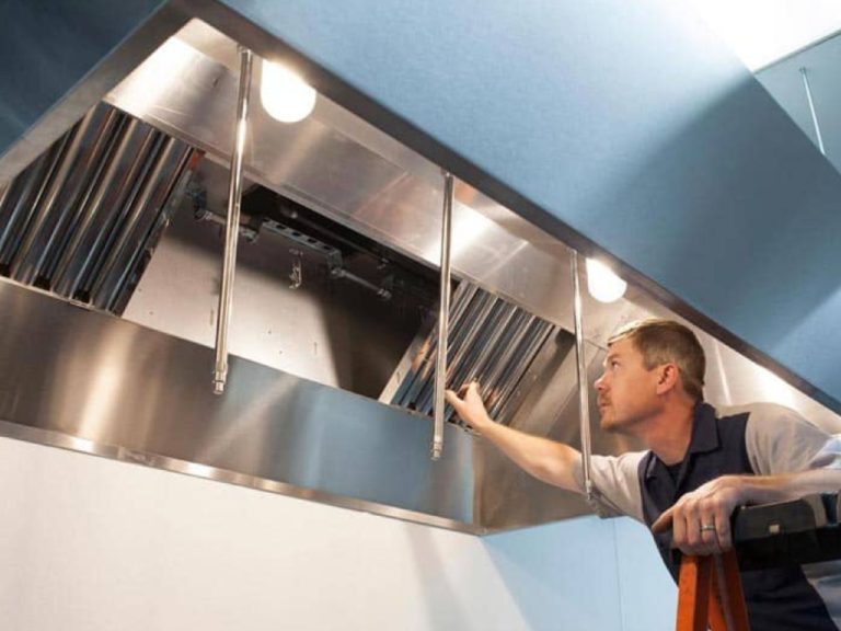 A man carefully installs a shiny stainless steel exhaust hood in a kitchen, focused on his task with tools nearby.