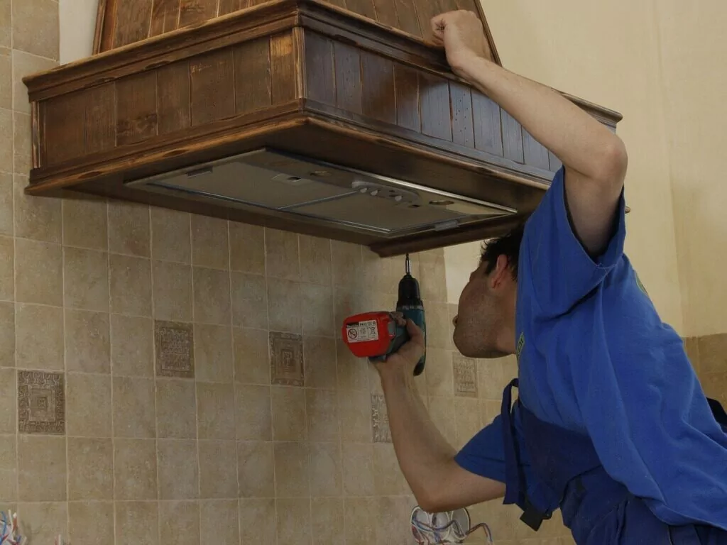 A man in a blue shirt operates a drill while installing a wood stove, focused on his task in a cozy setting.